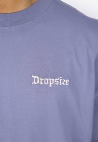 Lyseblå T-skjorte med rund hals i myk stoff, med hvit brodert tekst "Dropsize" på venstre bryst.