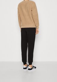 Felpa beige pelosa, pantaloni neri e sneakers nere con strisce bianche e logo, viste da dietro su uno sfondo semplice.