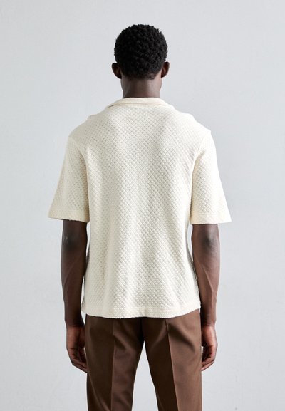 Korte mouwen, getextureerde top in off-white met een ruitpatroon, met een relaxte fit en een gebogen zoom, gecombineerd met bruine broeken.