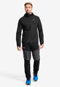 Svart zip-up jacka i texturerad tyg, kombinerad med svarta och grå byxor med fickor och reflektoraccenter. Kläderna fulländas av klara blå skor.