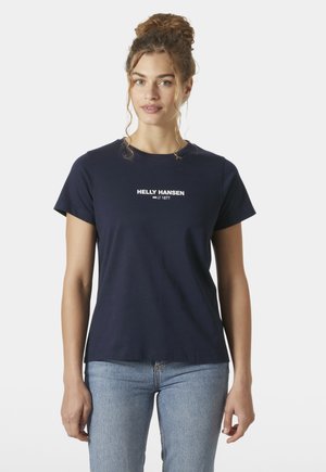 Vrouw met krullend haar in een knot, gekleed in een marineblauw Helly Hansen T-shirt en lichtblauwe spijkerbroek, tegen een effen witte achtergrond.