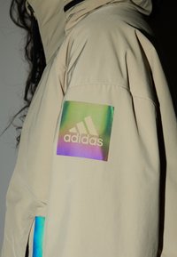 Beżowa kurtka z odblaskową, holograficzną naszywką logo Adidas na lewym rękawie. Gładka tekstura i luźny krój.