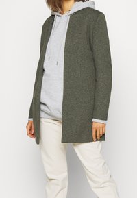 Olivengroen wollen blendjas met een rechte snit, met kraagontwerp en twee zijzakken, gedragen over een grijze hoodie.