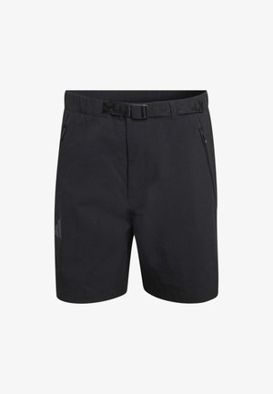 Mann trägt schwarze Sportshorts, weiße Kniestrümpfe, blaue und schwarze Laufschuhe, steht vor hellem Hintergrund.