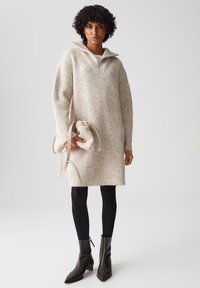 Vestito in maglia beige a costine con colletto con zip, abbinato a collant neri e stivaletti neri a punta. Porta una borsa morbida di un leggero beige.
