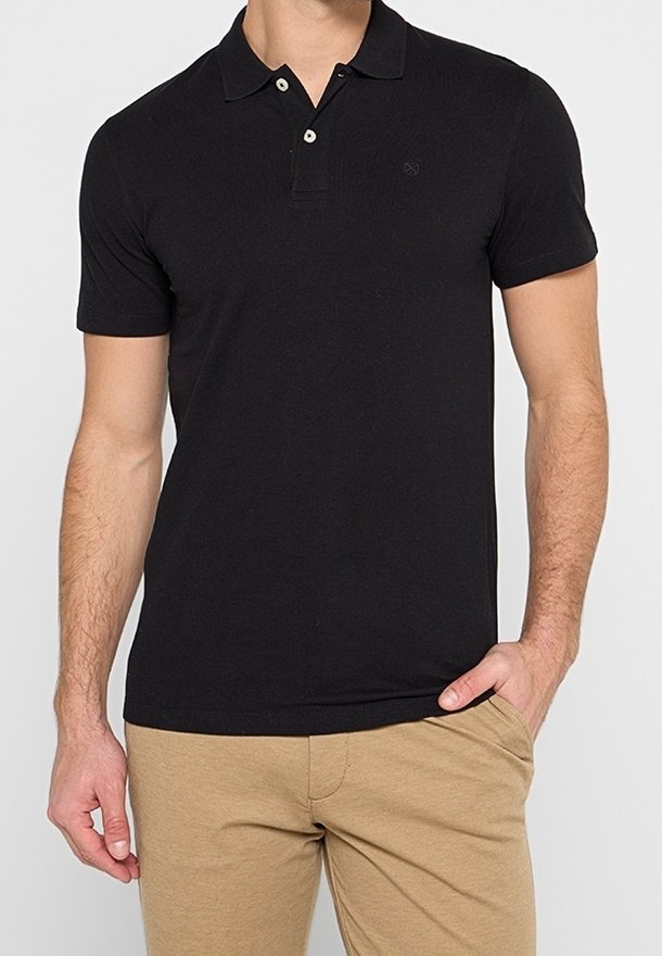 Homme portant un polo noir à manches courtes et un pantalon beige, avec une main dans la poche, sur un fond clair uni.