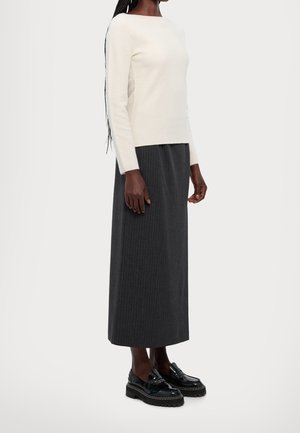 Femme portant un haut à manches longues crème, une jupe midi à fines rayures gris foncé et des mocassins à plateforme noirs, debout devant un arrière-plan uni.