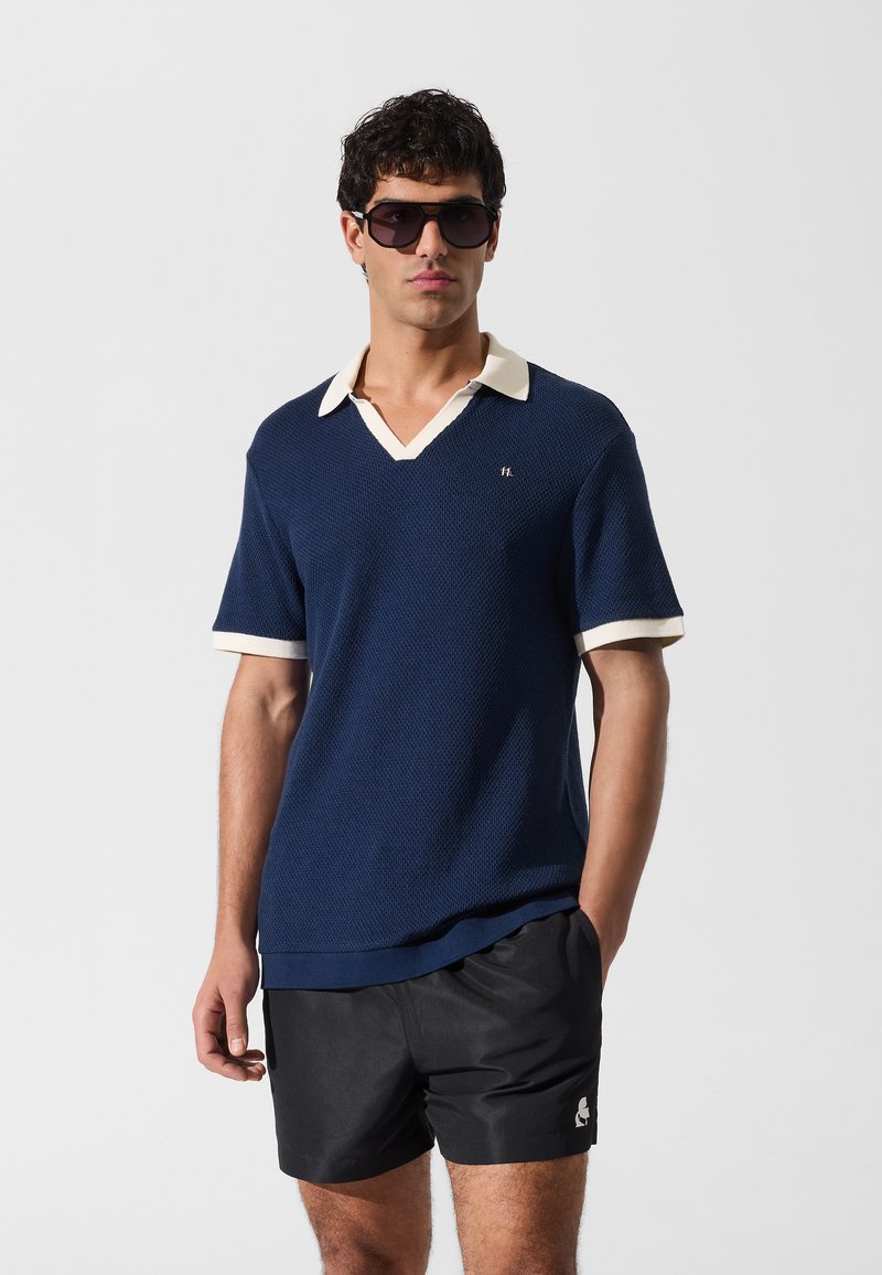 Polo texturé bleu marine avec col en V et col crème. Porté avec un short noir, arborant un petit logo sur le côté. Lunettes de soleil noires.