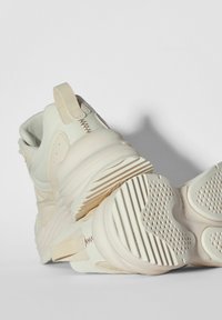 Beżowe sneakersy z teksturowanej tkaniny, masywną podeszwą i przeszytymi akcentami. Posiadają tylną pętelkę do zakupu i rowkowaną gumową podeszwę.