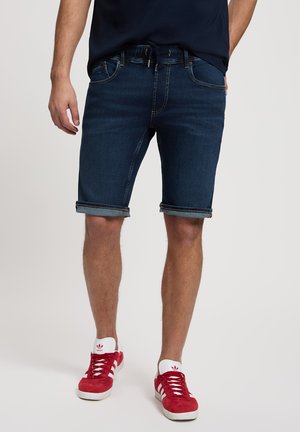 Herren Denim-Shorts, dunkelblau, mit taillierter Passform, gerollten Säumen, fünf Taschen und einem Kordelzug am Bund. Kombiniert mit roten Sneakers.