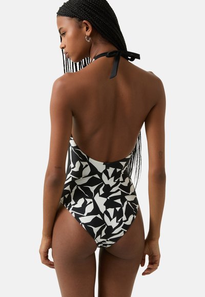 Maillot de bain une pièce à motifs noir et blanc avec un dos plongeant, un lien autour du cou et des jambes hautes. Fabriqué en tissu lisse et élastique.