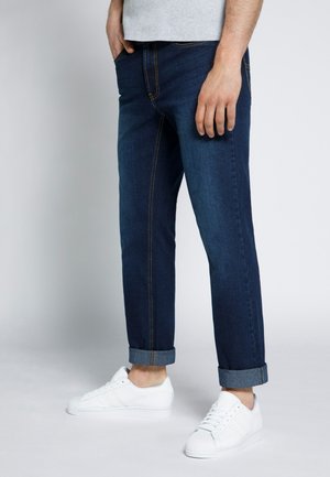 Donkerblauwe jeans met een rechte pasvorm, voorzien van zichtbare gele stiknaden, omgeslagen boorden en gecombineerd met witte sneakers.