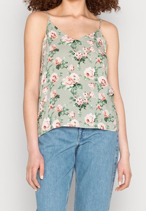 Frau trägt ein hellgrünes ärmelloses Oberteil mit pinken Rosen und blaue Jeans, steht vor einem einfarbigen Hintergrund.