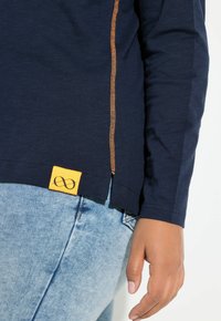 Camicia a maniche lunghe blu navy con dettagli di cuciture arancioni e un'etichetta gialla con il simbolo dell'infinito, abbinata a jeans di denim azzurro.