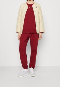 Chaqueta acolchada beige con cierre a presión, sudadera roja, pantalones rojos a juego y tenis blancos con acentos en gris claro. Logotipos de Nike en la ropa.