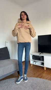 Beżowa bluza z kapturem, z kieszenią z przodu, połączona z obcisłymi niebieskimi dżinsami i wzorzystymi niebieskimi tenisówkami. W tle telewizor i grzejnik.