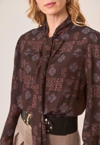 Blusa marrón con estampado, con cuello atado, que presenta diseños en azul y naranja quemado. La tela tiene una textura suave. Combinada con un cinturón negro.