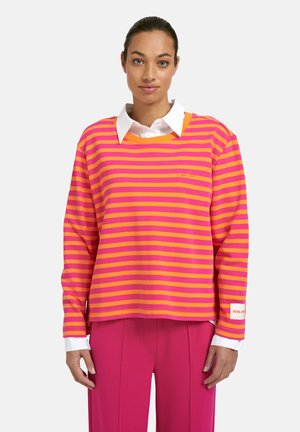 Woman wearing a bright pink and orange striped long-sleeve top over a white collared shirt and solid hot pink pants, standing against a white background.