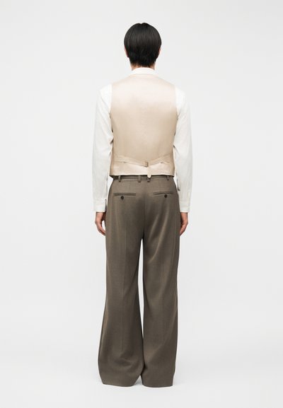 Άτομο που φοράει μπεζ γιλέκο πάνω από λευκό πουκάμισο με μακριά μανίκια και φαρδιά γκρι παντελόνια, στέκεται γυρισμένο με την πλάτη σε απλό φόντο.