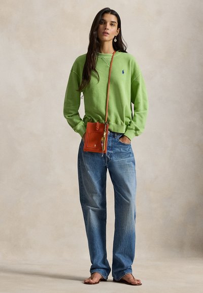 Kvinde stående barfodet iført en lysegrøn sweatshirt, løse blå jeans og en orange crossbody taske med en perleaccessory.