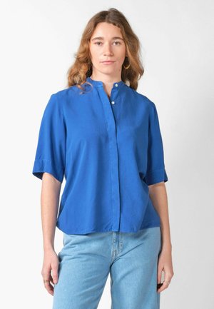 Mujer con cabello ondulado, pendientes de aro, camisa azul de manga corta con botones y vaqueros azul claro, de pie contra un fondo blanco.