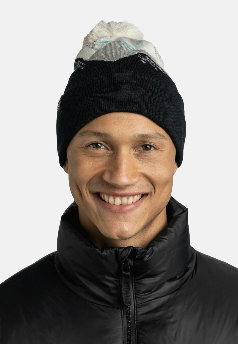 Gorro de punto negro con parte superior de patrón claro y pompón. Diseñado con un ajuste ceñido; se lleva con una chaqueta negra de cuello alto.