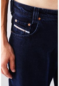 Dunkelblaue Jeans mit kupferfarbenen Nieten, einer kontrastierenden Tascheninnenfutter mit Karomuster und einer schmalen Passform. Auffällige Textur und Nähdetails.