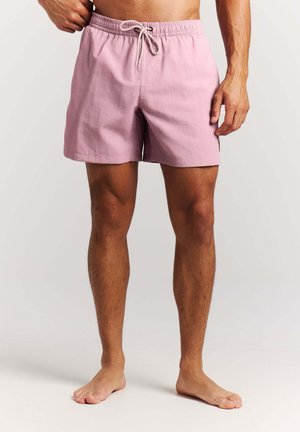 Mann trägt hellrosa Badehose mit elastischem Bund und Kordelzug, steht barfuß vor einem schlichten Hintergrund.