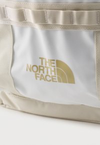 Κοντινό πλάνο λευκής υφασμάτινης τσάντας με μπεζ ιμάντες που φέρει χρυσό λογότυπο "The North Face" στην μπροστινή όψη.