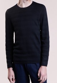 Pull noir à manches longues avec un motif texturé, col rond et coupe ajustée. Porté avec un pantalon bleu foncé, mettant en valeur les couleurs contrastées.