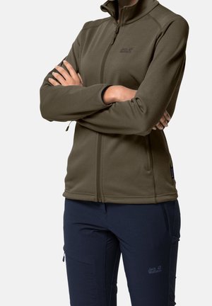 Persona che indossa una giacca verde oliva con zip e pantaloni outdoor blu scuro con loghi, braccia incrociate, in piedi davanti a uno sfondo semplice.