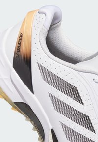 Valkoinen urheilukenkä mustilla verkkokoristeilla, kiiltävällä mustalla kannalla ja kultaisella liukuvärjäysyksityiskohdalla. Kenkässä on teksturoidut rei'itykset ja tyylikäs muotoilu.