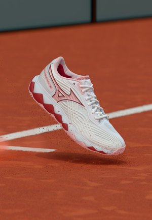 Chaussure de sport blanche et rose soulevant de la terre battue rouge sur un court de tennis près des lignes blanches de délimitation.