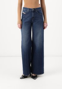 Jean en denim à jambes larges couleur bleu foncé avec une taille haute, dotés de poches avant et d'une fermeture par bouton. L'ourlet présente un discret détail de couture.