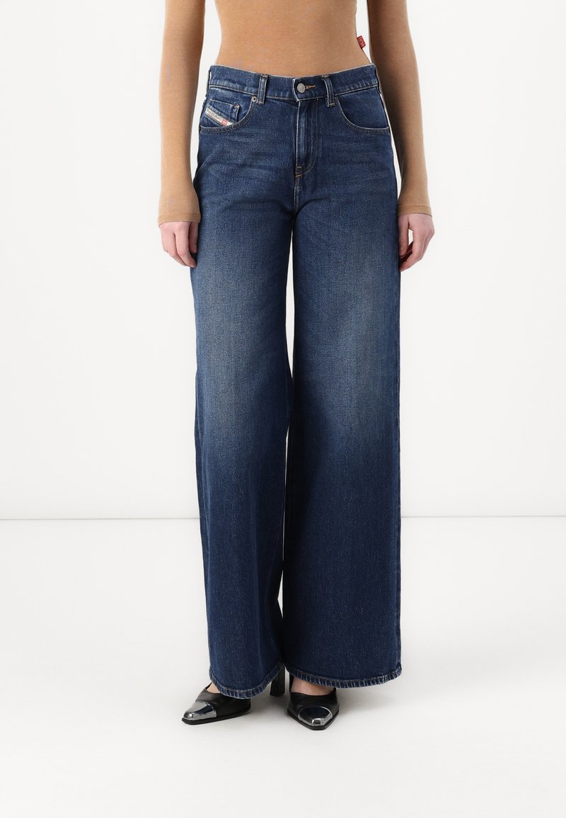 Jean en denim à jambes larges couleur bleu foncé avec une taille haute, dotés de poches avant et d'une fermeture par bouton. L'ourlet présente un discret détail de couture.