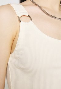 Gros plan d'une personne portant un haut sans manches crème côtelé avec un détail anneau en métal sur l'épaule et un collier chaînette argenté.