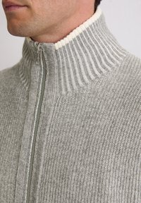 Grauer Strickpullover mit hohem Kragen, versehen mit einem kremfarbenen Rippbesatz und einem Reißverschluss vorne, der aus einem strukturierten, leichten Material besteht.