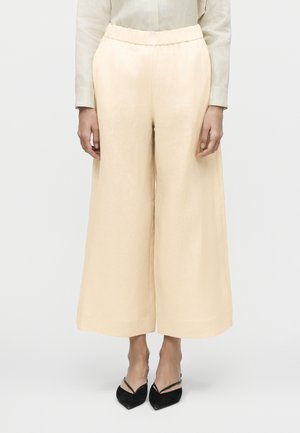 Kvinde iført brede cremefarvede bukser kombineret med sorte spidse slingback-sko, stående mod en ensfarvet baggrund.