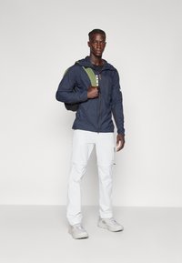 Marineblauwe lichte jas met capuchon, ritssluiting aan de voorkant en wit logo, gecombineerd met witte cargobroek en grijze sneakers. Groene rugzak op de schouder.