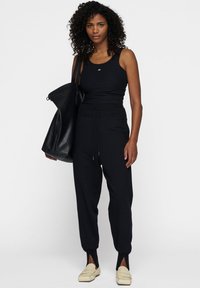 Top sin mangas negro con textura de canalé, combinado con pantalones jogger negros que tienen una cintura con cordón y puños con abertura, complementado con un bolso grande negro.