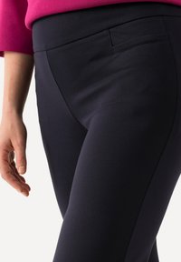 Pantalones elásticos azul marino con una textura suave, cintura de altura media y un pequeño bolsillo lateral, parcialmente mostrados con una mano descansando sobre la pierna.