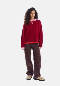 Czerwony, oversize'owy sweter z kontrastującym różowym kołnierzem, zestawiony z brązowymi spodniami o szerokich nogawkach i beżowymi butami trekkingowymi. Widoczne kręcone włosy i dodatki.