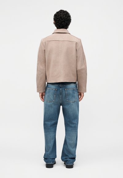 Άτομο με σγουρά μαλλιά, ντυμένο με μπεζ μπουφάν και φαρδιά μπλε τζιν, στέκεται με την πλάτη σε έναν απλό φωτεινό φόντο.