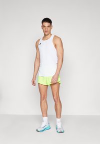 Witte tanktop met zwart logo, neon groene shorts en lichtblauwe sportschoenen. De stof lijkt lichtgewicht en geschikt voor sportactiviteiten.