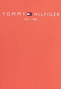Korallowa bawełniana koszulka z krótkim rękawem, z białym i granatowym logo "TOMMY HILFIGER" z napisem "EST. 1985" poniżej, gładka w dotyku.