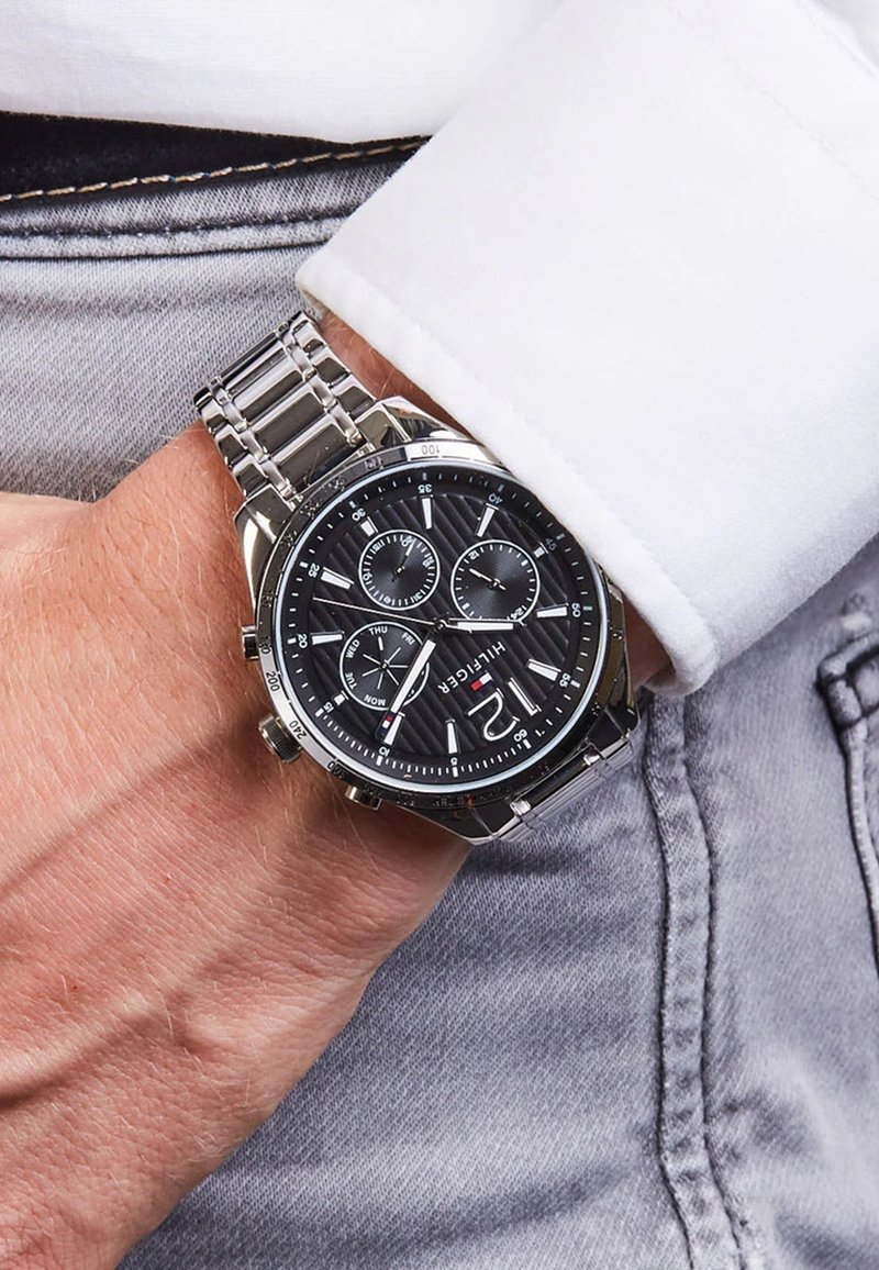 Montre en acier inoxydable argent�é avec un cadran texturé noir, trois sous-cadrans, aiguilles argentées et un bracelet métallique à maillons. Portée au poignet.