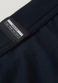 Tmavě modrá tkanina s hladkým povrchem. Na švu je přišitá černá nálepka s bílým textem, která uvádí značku a pokyny pro údržbu.