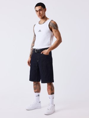 Jeune homme avec des tatouages portant un haut blanc sans manches, un short noir mi-long, des chaussettes blanches et des baskets blanches, debout devant un fond uni.