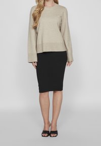 Beige Strickpullover mit langen Ärmeln, kombiniert mit einem schwarzen, figurbetonten Bleistiftrock. Trägt dazu schwarze Sandalen mit offenem Zeh. Glatte Texturen überall.