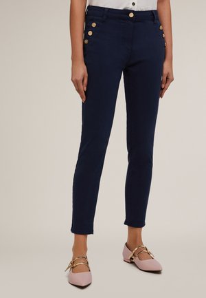 Pantalons skinny bleu marine avec des accents de boutons dorés sur les côtés, présentant une texture lisse et une coupe ajustée. Portés avec des ballerines rose pâle.
